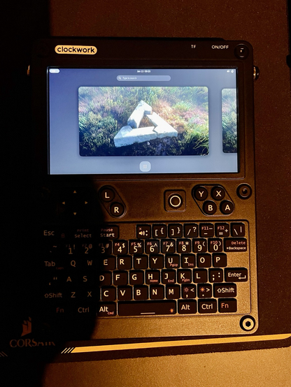 Photo of a ClockworkPi uConsole with GNOME desktop environment visible on screen. The postmarketOS grass wallpaper is visible on the screen