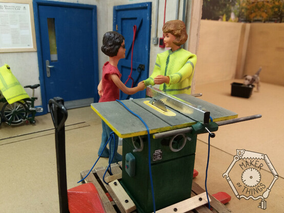 The scene inside a 1:12 scale workshop. In the background are two blue doors, a Health and Safety at work act poster, a folded Brompton bicycle, a red dog lead hanging up, and an openroller shutter showing an enclosed yard outside the workshop.
There is also narrow gauge tram tracks set into the workshop floor.
The view outside is a work in progress.

Inside the workshop there are two Caucasian women doll figures. Harriet has dark hair, glasses, and is wearing a red tee shirt and blue jeans, Daisy has fair air and is wearing a grubby high viz coat. They are shaking hands.

In front of them is a large Wadkin AGS10 table saw painted green. It is on a wooden pallet and tied down with rope. The pallet is on an orange pallet truck having just been brought into the workshop through the roller shutter.

Outside in the yard is a grey and black needle felted mongrel dog called Monty Dog, and he is trying to pick up a black recycling collection box.

In the bottom right hand corner of the image is a watermark showing the Maker of Things logo consisting of an outline of a circular saw blade surrounded by a spanner, a hand saw, a ruler, a screwdriver, a cable and plug, and a pencil forming a hexagon. Inside the tools are the words Maker of Things.