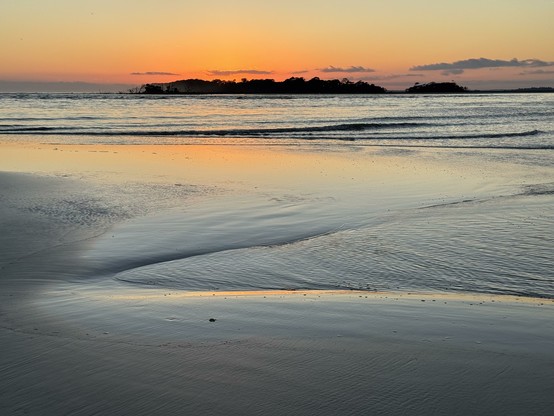 Vivid orange and blue, sand water and sky, looking across an inlet to a dark island in the distance.