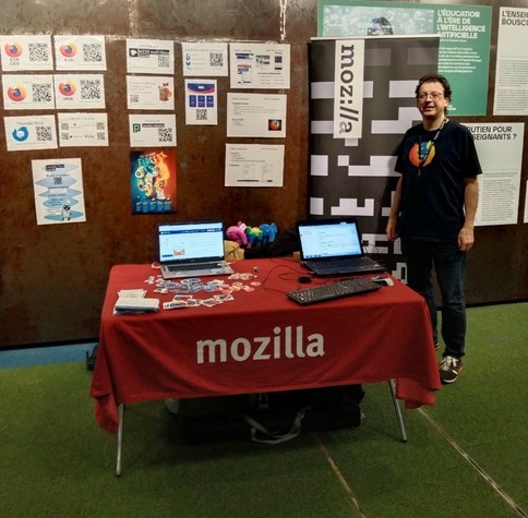 Mozillien Hellosct1 devant un kakémono Mozilla, à côté d’un stand Mozilla avec dessus deux ordinateurs portables, des stickers et des éléphants en peluche ; sur le mur derrière sont affichées diverses affiches dont certaines arborent un code QR.