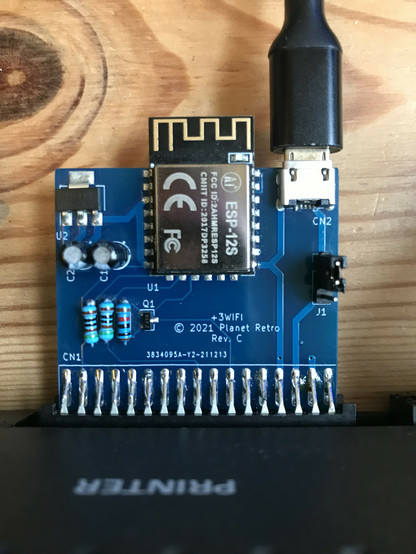 Blue PCB plugged in to the printer port of a ZX Spectrum +3. The PCB has an ESP-12 WiFi module on it, and a USB cable plugged in to power it.