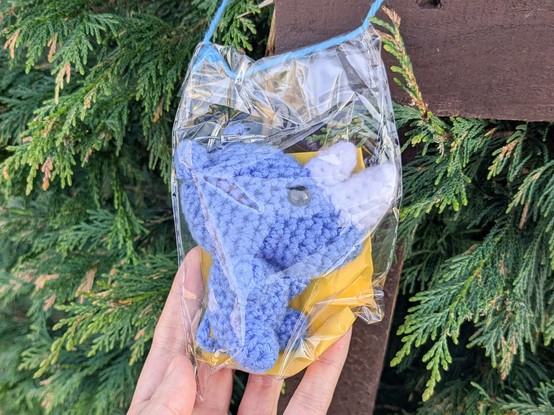 A small blue crocheted rhinoceros in a plastic bag, haging from a bit of fence in among the leaves of a bush. There's a piece of yellow paper in the bag too but it's almost completely obscured.