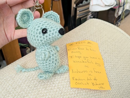 A light greeny-blue crocheted mouse with black nose and eyes and a keychain attached to its head. It's sitting on the arm of my couch, next to a handwritten note on yellow paper that reads "I am free to take away, I hope you have a wonderful day. Kindness is free. Random Act of Crochet Kindness UK"