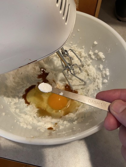 a spoon of powdered salt with the spoon labelled “DASH” or 1/8th teaspoon, held over a bowl of otherwise wet ingredients