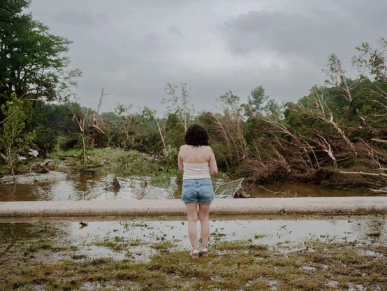 Seen from behind, a woman in denim shorts and white sleeveless tank top looks out at the watery devastation of the flood, with some home wreckage and rows of downed trees among the receding waters.  The sky is an ominous cloudy gray.