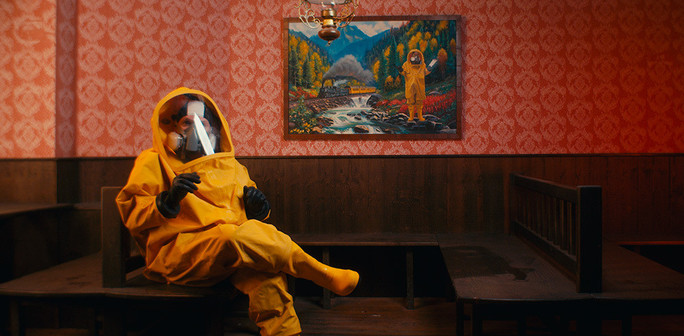 A person in a yellow hazmat suit sits in a dimly lit wooden booth against red patterned wallpaper. Above them hangs a painting showing a similar figure in a hazmat suit standing by a mountain stream, creating a surreal mirror effect. Still from the 2025 documentary Hacking at Leaves.