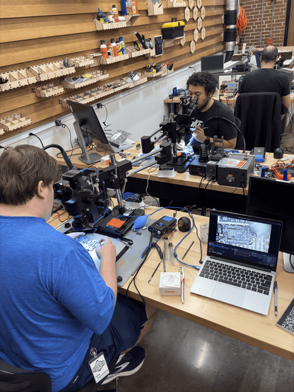 Students in a microsoldering workshop focus on circuit boards under microscopes, surrounded by laptops, precision tools, and organized wall-mounted supplies.