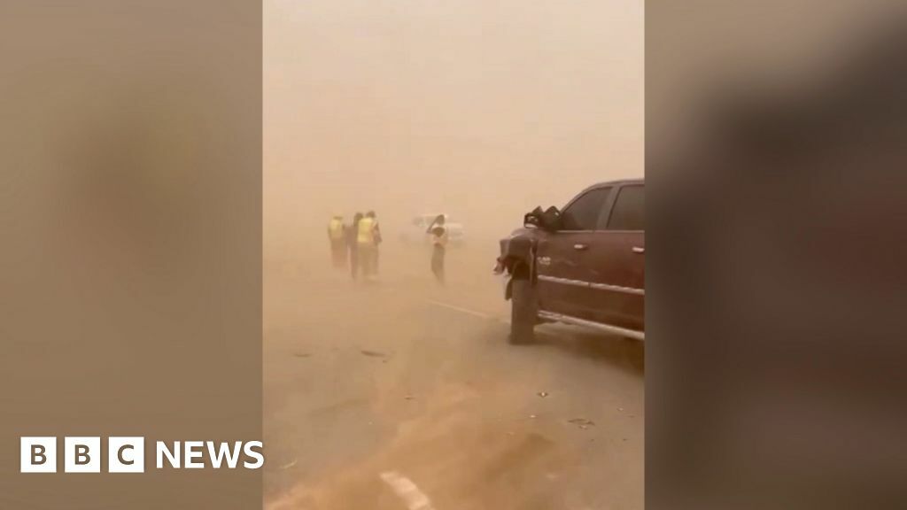 A maroon car sits on a dust-covered road immersed in a dust-cloud as people in high-vis jackets stand beyond it.